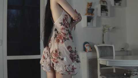 Media: Video of a slender woman with long black hair, wearing a sheer, floral-patterned nightgown, standing in a softly lit, minimalist room with white furniture and shelves filled with books and toys.