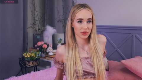 Media: Video of a blonde woman with fair skin, wearing a pink off-shoulder top, sitting on a pink bed. Background features a floral arrangement, white feathers, and gray wallpaper.