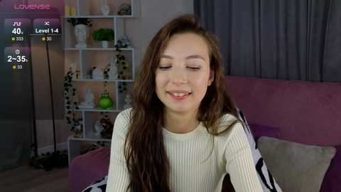 Media: Video of a young woman with long brown hair, fair skin, and a light knit sweater, smiling in a modern living room with shelves, plants, and a purple couch.