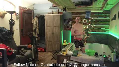 Media: Video of a messy living room with a man wearing glasses, no shirt, and black shorts, standing near a green-lit pool table.