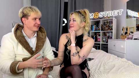 Media: Video of a blonde man and woman in a bedroom. The man wears a white robe, and the woman, with tattoos, dons black lingerie. They are smiling, sitting on a bed.