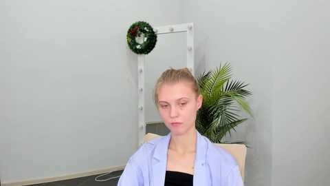 Media: Video of a young Caucasian woman with blonde hair, wearing a light blue button-up shirt over a black top, sitting indoors with a green wreath, a white Christmas tree, and a potted palm plant in the background.