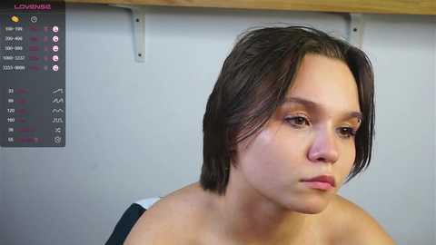 Media: Video of a young woman with short dark hair, fair skin, and a neutral expression, wearing a black tank top. Background shows a gray wall with a mounted wooden shelf.