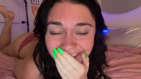 Media: A video of a young woman with long black hair, green nail polish, lying on a bed, smiling, covering her mouth, wearing a red bra, in a dimly lit bedroom.