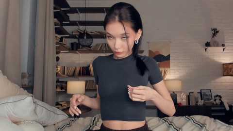 Media: A young Asian woman with long black hair tied back, wearing a black crop top, stands in a modern loft bedroom with exposed beams, shelves filled with books, and a cozy lamp.