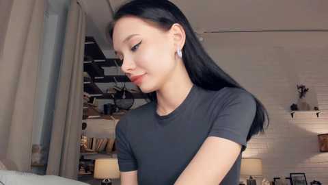 Video of an East Asian woman with long black hair, wearing a grey t-shirt, looking contemplative in a modern, cozy room with white brick walls, wooden shelves, and soft lighting.