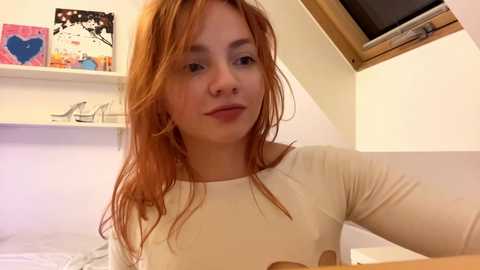 Media: Video of a fair-skinned redhead woman with straight hair, wearing a white long-sleeved shirt, sitting in a bedroom with a sloped ceiling.