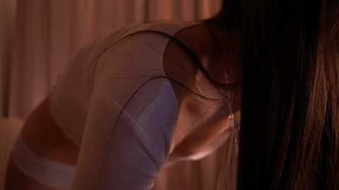 Media: Video of a woman with long dark hair, wearing a white tank top, leaning forward with her back arched, against a blurred, dimly lit background.