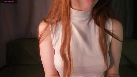 Video of a fair-skinned woman with long, straight, reddish-brown hair, wearing a sleeveless white turtleneck sweater, standing against a dimly lit, green couch in a room with dark curtains.