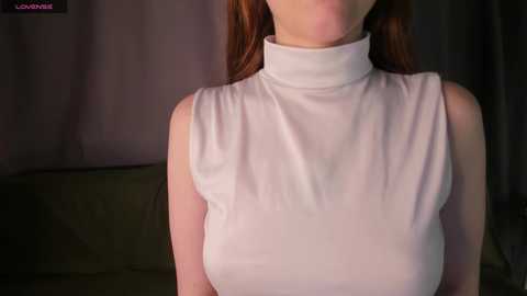 A video of a fair-skinned woman with long, straight auburn hair, wearing a sleeveless, high-neck, white turtleneck dress, sitting on a green couch in a dimly lit room.