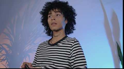 Media: Video of a young man with curly black hair, wearing a black-and-white striped shirt, standing against a blurred palm tree and blue sky background.