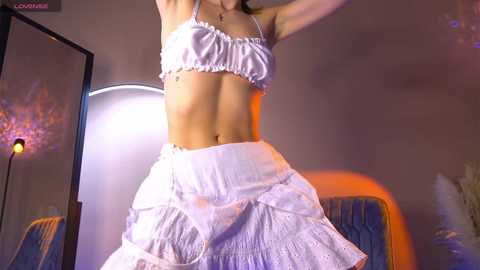 Media: Video of a slender, light-skinned woman with medium breasts, wearing a white frilly bra and matching high-waisted skirt, posing indoors with a mirror and glowing lights in the background.