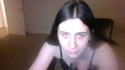 Media: A video shows a young woman with long black hair, green eyes, and pale skin, wearing a black bra, kneeling on a beige carpet in a dimly lit room with white walls and a door in the background.