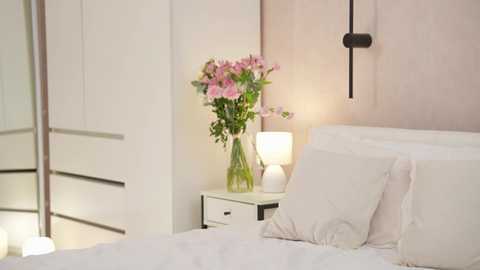 Media: Video of a minimalist bedroom featuring a white bed, white nightstand with a vase of pink flowers, and a wall-mounted lamp on a soft pink wall.