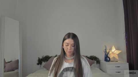 Media: A video of a young woman with long, straight brown hair, wearing a white graphic tee and pearl necklace, standing in a minimalist bedroom with a white bed, a blue star lamp, and a purple curtain.