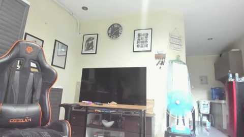 Media: A video of a modern, minimalist living room with beige walls, featuring a black gaming chair, flat-screen TV, wall art, and a blue air purifier.