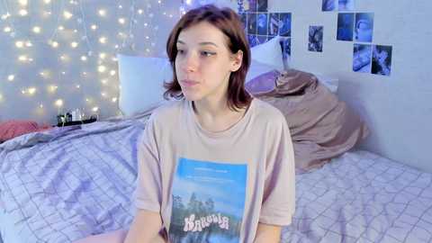 Media: Video of a young, fair-skinned woman with shoulder-length brown hair, wearing a loose beige t-shirt, sitting on a bed with a blue blanket, surrounded by string lights and photo frames.