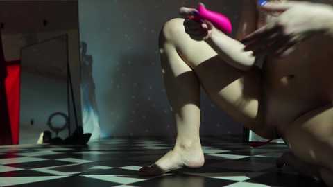 Media: Video of a nude, fair-skinned woman with large breasts, crouching on a black-and-white checkered floor. She holds a bright pink vibrator, casting a shadow on the wall. Background includes a red curtain and a mirror, creating a surreal, intimate ambiance.