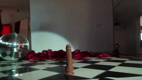 Media: Video of a wooden dildo on a black-and-white checkered chessboard, surrounded by red satin, in a dimly lit room with a shiny disco ball and a mirror.