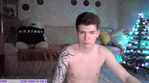 Media: Video of a shirtless, pale-skinned young man with short dark hair, sporting a detailed tattoo on his left arm, sitting indoors in a cozy living room with a Christmas tree and plush toys.