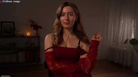 Media: Video of a smiling woman with light skin, long wavy brown hair, wearing a red lace dress and matching long gloves, sitting in a dimly lit room with a fireplace and bookshelves.