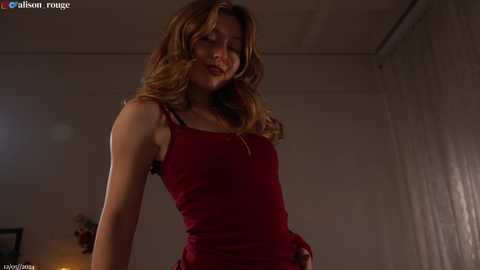 Media: Video of a slim, light-skinned woman with wavy brown hair, wearing a tight, sleeveless red dress, standing in a dimly lit room with a white curtain background.