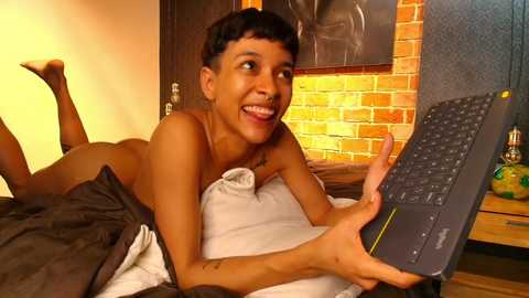 Media: Video of a smiling, topless woman with short hair, lying on a bed with a laptop, in a cozy room with brick wall and warm lighting.