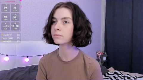 Video of a young woman with short, dark brown hair, wearing a beige shirt, standing in a bedroom with a white wall, a purple string of lights, and a dark curtain.