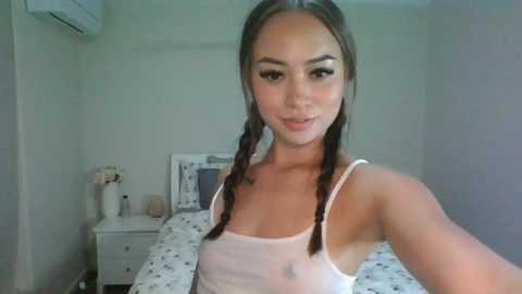 Media: Video of a young woman with light skin, long brown hair in braids, wearing a white tank top, smiling, standing in a simple bedroom with white walls, bed, and nightstand.