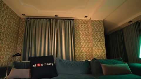 Media: A video of a dimly-lit living room with patterned wallpaper, teal couch, and a \"Mostbet\" pillow, featuring a TV screen in the background.