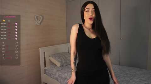 Media: A video of a woman with long dark hair, fair skin, and a slender build, wearing a tight black dress, standing in a modern bedroom with a grey bedspread.