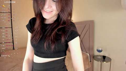 Media: Video of a young, fair-skinned woman with long, dark brown hair, wearing a black crop top and high-waisted black shorts, smiling in a dimly lit bedroom with a bed and a glass of wine on a side table.