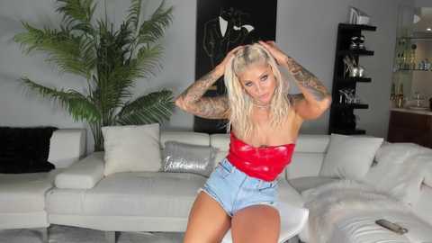 Media: Video of a tattooed blonde woman in a red, strapless top and denim shorts, posing in a modern, minimalist living room with white furniture and black accents.