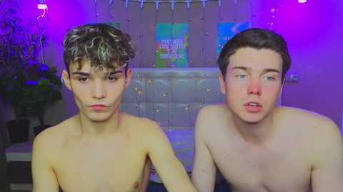 Media: Video of two shirtless young men with fair skin, one with curly brown hair, the other with short brown hair, looking serious and relaxed in a dimly lit room with purple lights, posters, and a plant in the background.