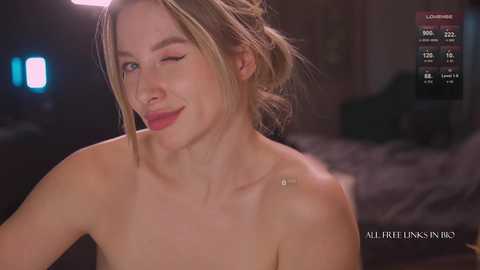 Media: A video of a topless young woman with fair skin, blonde hair, and blue eyes, smiling with a wink, set in a dimly lit bedroom.