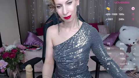 Media: Video of a fair-skinned woman with blonde hair in a pink room, wearing a glittery silver one-shoulder dress, sitting on a chair.