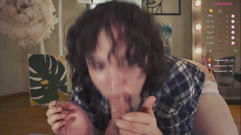 Media: A blurred video of a woman with messy black hair performing oral sex on a man, wearing a plaid shirt and white panties. Background shows a green and white plant and a wooden floor.