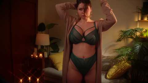 Media: Video of a curvy, fair-skinned woman with dark hair, wearing teal lingerie and a beige cardigan, standing in a dimly lit bedroom with warm lighting, potted plants, and a bed with yellow pillows.