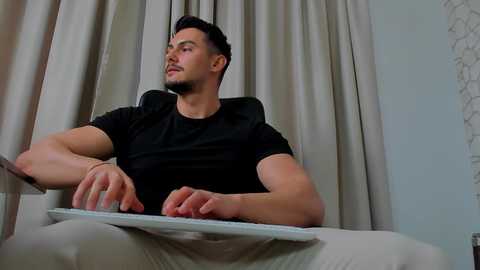 Media: Video of a fit, light-skinned man with short dark hair, wearing a black T-shirt and beige pants, sitting on a couch against beige curtains and a white wall, holding a remote.