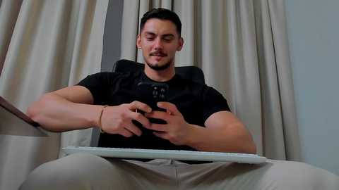 Media: Video of a muscular, fair-skinned man with short dark hair and a beard, wearing a black shirt and gray pants, sitting on a modern chair, playing a video game.