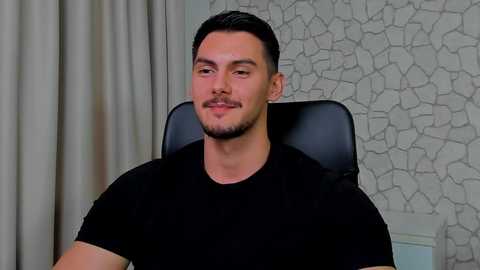 Media: Video of a man with short, dark hair and a trimmed beard, wearing a black T-shirt, seated in a black leather chair, with a textured, gray-patterned wallpaper and white curtains in the background.