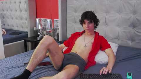 Media: Video of a young, slim, shirtless man with curly hair, wearing only gray briefs, sitting on a bed with a red wall and tufted gray headboard.