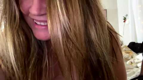 Media: A close-up video of a smiling woman with long, messy blonde hair, partially covering her face, taken indoors in a softly lit room with a bed and a plant in the background.