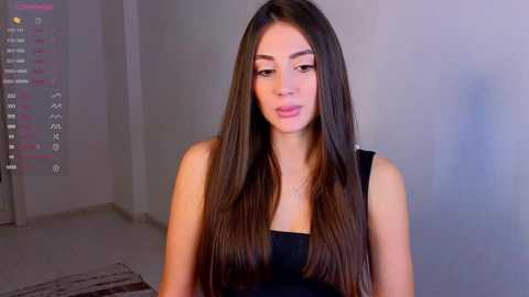 Media: Video of a young woman with long, straight brown hair, wearing a black top, standing in a dimly lit room with a white wall and a window.