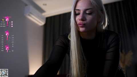 Media: Video of a blonde woman in a black top, making a pouty face, in a dimly lit room with a projector displaying colorful abstract art on a wall.