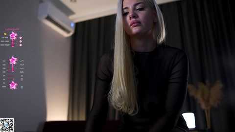 Media: Video of a blonde woman with long hair, wearing a black sheer top, standing in a dimly-lit room with dark curtains, air conditioner, and a projector screen displaying colorful graphics.