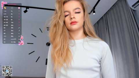 Media: A video of a young, fair-skinned woman with long, wavy blonde hair, wearing a light grey long-sleeve top, standing in a minimalist room with grey curtains and a black wall clock.