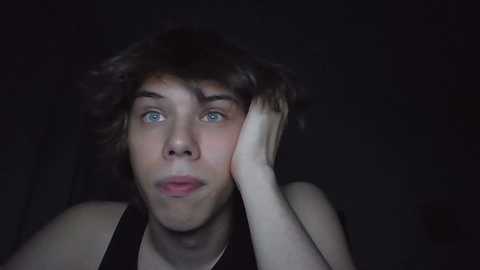 Media: Video of a young man with shoulder-length brown hair and blue eyes, resting his head on his hand, wearing a black tank top, against a dark background.