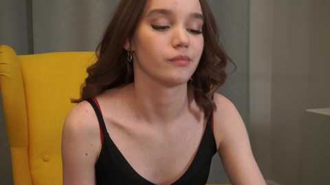 A video of a young woman with fair skin and long brown hair, wearing a black tank top with red straps, sitting on a yellow chair.