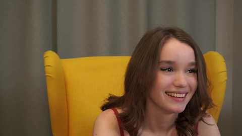 Video of a young woman with light skin and wavy brown hair, smiling, wearing a red top, sitting in a mustard-yellow armchair against a gray curtain background.